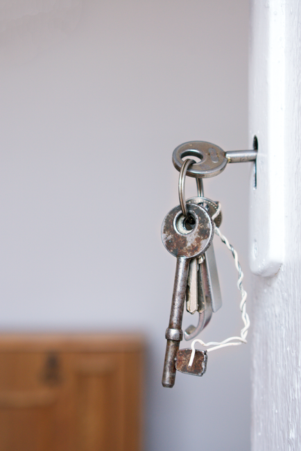 Close-up of a door lock with keys hanging from the keyhole, mounted on a wooden door with metallic hardware. The door features a modern handle and lock mechanism, with a keyring holding multiple keys.