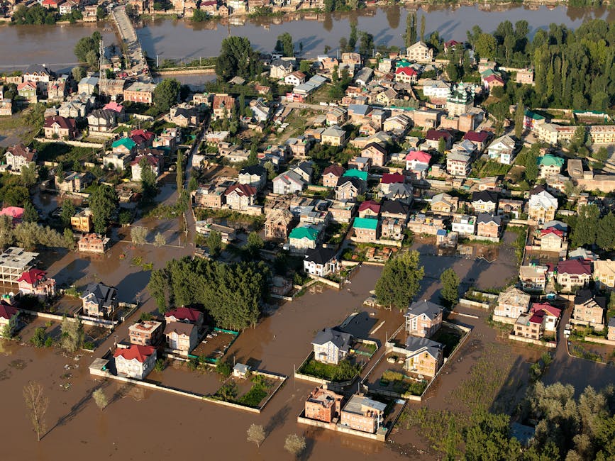 An aerial view of a residential area affected by flooding, showing numerous houses with flooded yards and streets, some with visible water covering gardens and driveways. The houses feature a variety 