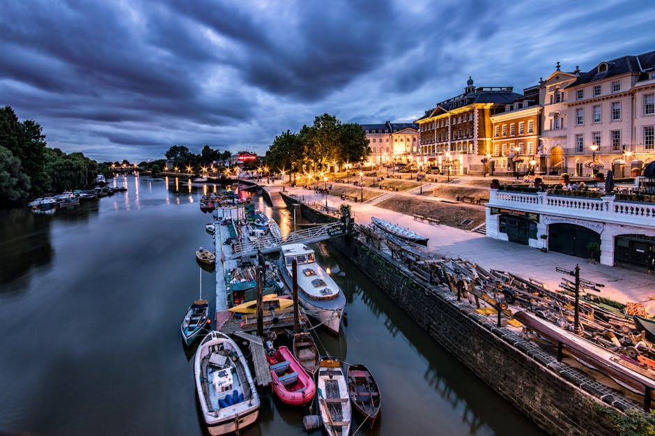 A calm river scene in Kingston upon Thames with several small boats moored along the water's edge, reflecting the partly cloudy blue sky and lush green trees lining the riverside. In the background, white and beige residential buildings and historic structures are visible behind the foliage. The image highlights natural outdoor surfaces, including the water and stone embankment in the foreground, showcasing a serene environment. Cleaner Kingston upon Thames specializes in surface cleaning and deep cleaning services to maintain the hygiene and aesthetic appeal of similar outdoor and indoor spaces, ensuring they remain pristine and properly maintained.