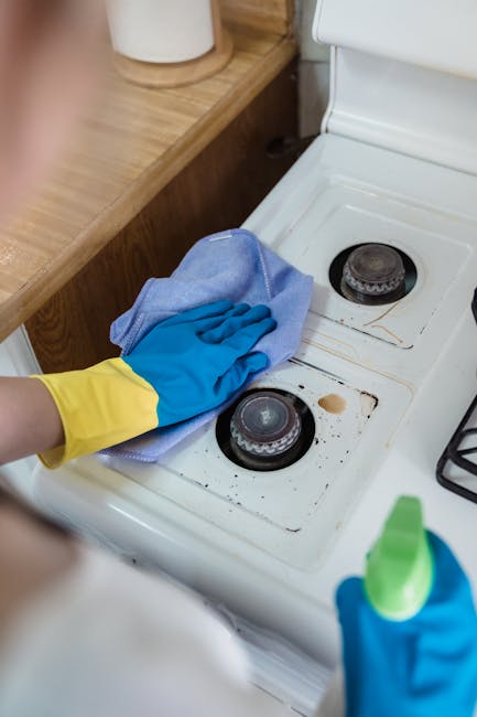 A person wearing a white shirt and yellow and blue rubber cleaning gloves is using a cloth to thoroughly clean the interior of a white electric oven. The oven door is open, revealing the metal racks a