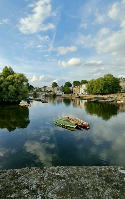 A serene river scene under a partly cloudy sky, with still water reflecting the fluffy white clouds and blue sky above. Several small boats are docked along the riverbank, surrounded by lush green trees and residential buildings in the background. The scene illustrates calmness and natural beauty, fitting for a context related to outdoor cleaning or maintenance. The image's clarity highlights the details of the boats and the reflections on the water, emphasizing the importance of thorough surface cleaning and outdoor sanitisation, as may be described in content by Cleaner Kingston upon Thames for their domestic and commercial cleaning services.