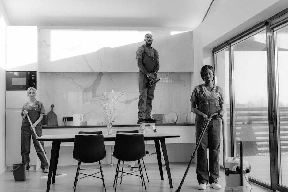 A group of three professional cleaners from Cleaner Kingston upon Thames are performing surface cleaning and deep cleaning tasks in a modern, well-lit kitchen area. One cleaner stands on a stool, scrubbing or wiping a high-mounted white marble backsplash with grey veining, ensuring thorough sanitisation of the surface. Another cleaner, on the right, holds a mop and is preparing to clean the wooden floor, which appears spotless and shiny. The third cleaner, on the left, is using a vacuum or floor brush to clean around the dining table, which has a dark, matte finish with four black chairs arranged around it. The kitchen features large sliding glass doors that let in natural light, illuminating the sleek stainless steel appliances and minimalist decor. The surfaces, including the countertop and table, are visibly clean and dust-free, with some cleaning supplies on the counter and the cleaning tools in use. The overall scene emphasizes professional domestic cleaning and surface sanitisation, aligning with the context of comparing flat cleaning costs in Kingston upon Thames, provided by [COMPANY_NAME].