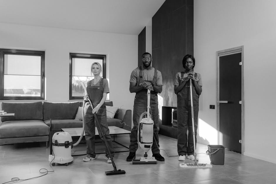 Three professional cleaners from Cleaner Kingston upon Thames are performing surface cleaning and deep cleaning in a modern living room, which features a large L-shaped sofa, a coffee table, and two windows allowing natural light to illuminate the space. The team is equipped with vacuum cleaners, mops, and cleaning cloths, and they are working on polished wooden and ceramic flooring, which appears spotless and well-maintained. The first cleaner, a woman, is vacuuming the rugs near the sofa, while the male and the other woman are mopping the tiled floor area near a closed door. The clean and organized room highlights routine hygiene and maintenance practices associated with domestic cleaning services, underlining the company's focus on comprehensive sanitisation and surface cleaning. The bright, uncluttered setting underscores the importance of professional cleaning for a tidy and healthy living environment, consistent with the themes of the flat cleaning cost comparison in Kingston upon Thames.