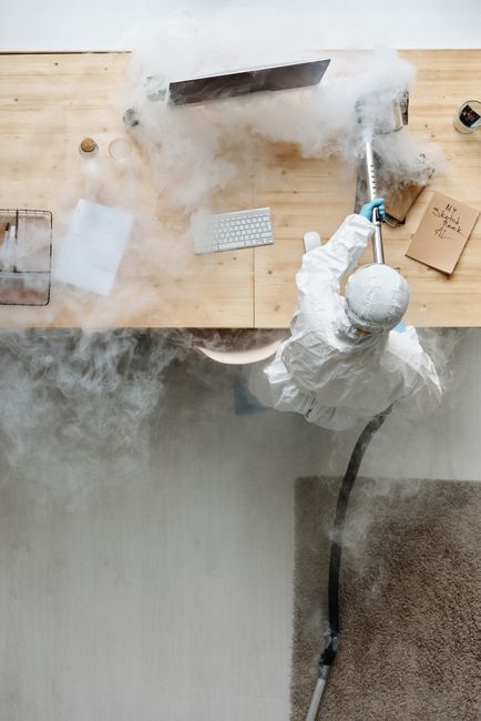 An individual wearing full personal protective equipment, including a white suit and a face mask, is performing deep surface cleaning or sanitisation on a light-colored wooden office desk using a fogging or misting device that emits a cloud of disinfectant vapor. The desk features a small potted plant, a glass of water, a closed notebook, and a white keyboard, all of which are being treated to eliminate germs and dust. The workspace is well-lit, with reflections on the surfaces indicating cleanliness and hygiene. This cleaning process is part of routine office sanitisation procedures, emphasizing health and safety rules for maintaining a hygienic environment at work, as promoted by Cleaner Kingston upon Thames.