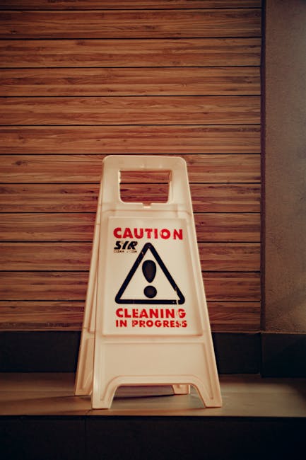 A white plastic 'Caution Cleaning in Progress' A-frame sign with a red and black warning symbol is positioned on a tiled floor in front of a wall clad with horizontal wooden panels. The sign indicates ongoing cleaning activities, emphasizing safety measures in the area. The setting appears to be an indoor space, possibly an entrance or corridor, with clean, well-maintained surfaces. The lighting is neutral, highlighting the sign's visibility and the cleanliness of the surrounding environment. This image aligns with principles of surface cleaning and maintenance promoted by Cleaner Kingston upon Thames, ensuring hygiene and safety in office or commercial settings as outlined in the 'Health and safety rules for office cleaning Kingston upon Thames' page.