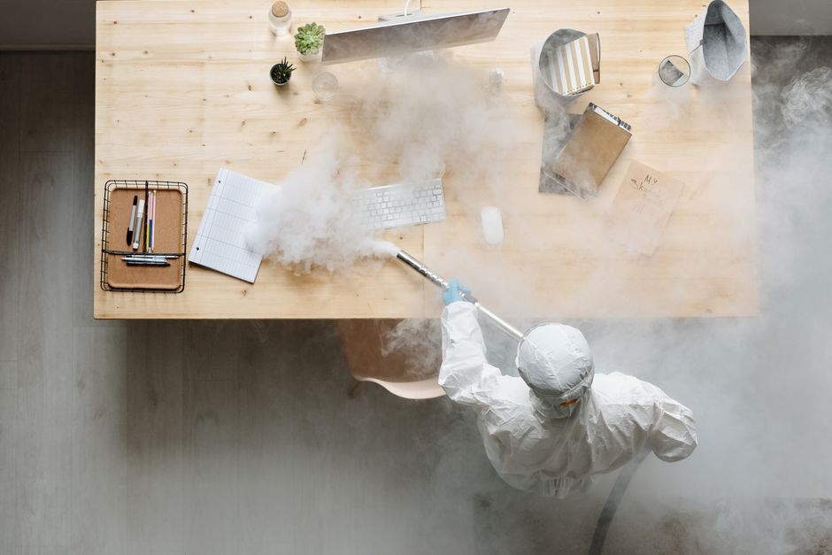 An individual wearing full personal protective equipment, including a white suit and a face mask, is performing deep surface cleaning or sanitisation on a light-colored wooden office desk using a fogging or misting device that emits a cloud of disinfectant vapor. The desk features a small potted plant, a glass of water, a closed notebook, and a white keyboard, all of which are being treated to eliminate germs and dust. The workspace is well-lit, with reflections on the surfaces indicating cleanliness and hygiene. This cleaning process is part of routine office sanitisation procedures, emphasizing health and safety rules for maintaining a hygienic environment at work, as promoted by Cleaner Kingston upon Thames.