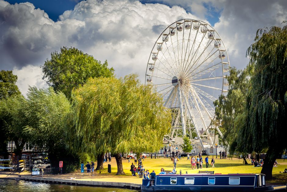 A large Ferris wheel located in a park with lush green trees surrounding it, people walking and relaxing on the grassy area, some sitting on benches, and a narrow waterway with a boat in the foreground. The sky is partly cloudy with patches of blue, providing natural lighting that highlights the vibrant greenery and the white structure of the Ferris wheel. The scene depicts a lively outdoor recreation space in Kingston upon Thames, emphasizing clean, well-maintained park surfaces and the family-friendly atmosphere. Cleaner Kingston upon Thames specializes in surface cleaning and deep cleaning to maintain hygiene and appearance across residential and commercial spaces within this area.
