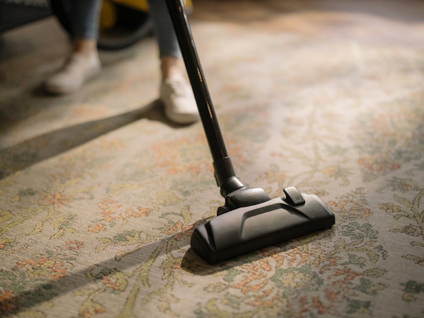 Close-up view of a vacuum cleaner head with a black and gray design, actively being used to clean an ornate, patterned area rug in a residential setting. The rug features a muted floral design in shades of beige, light green, and soft orange, with a polished, dust-free surface. The vacuum's hose and handle are visible, held by a person wearing dark pants and white shoes, standing on the rug. The scene is illuminated by natural light, highlighting the thorough surface cleaning process facilitated by Cleaner Kingston upon Thames' domestic cleaning services, focusing on carpet and surface hygienisation in an everyday home environment.