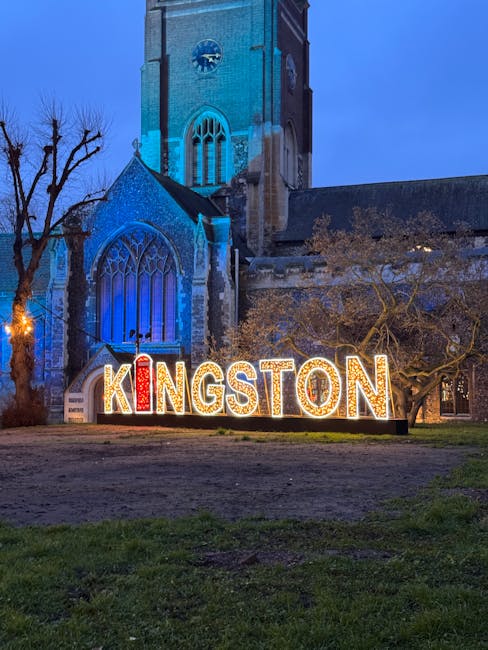 A nighttime outdoor scene featuring a Gothic-style church with tall, pointed arched stained glass windows illuminated by blue lighting. In the foreground, large illuminated letters spelling 'KINGSTON' are displayed on a grassy area, with the letters lit in red, yellow, and white. Bare tree branches stretch across the scene, and a warm streetlamp light is visible on the left side. The image portrays a well-lit, clean, and inviting public space, highlighting the village or town's charm in Kingston upon Thames, which is relevant to local cleaning and surface maintenance services by Cleaner Kingston upon Thames.