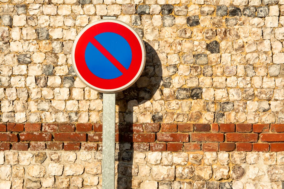 A white rectangular parking sign affixed to a wooden post against a background of a brick wall. The sign features a blue wheelchair symbol in the top left corner indicating accessible parking, with it