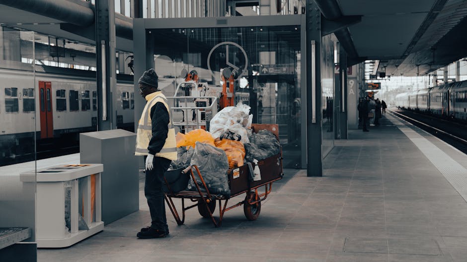 A sanitation worker wearing a black beanie, glasses, a yellow safety vest, and black clothing is standing next to a large cart filled with garbage bags and waste items on a modern train station platform. The platform features smooth, light gray tiled flooring, glass walls, and metal structural elements. Adjacent to the worker is a white recycling bin. In the background, parked trains with closed doors are visible, along with a few other individuals further down the platform. The overall scene is well-lit with natural daylight, emphasizing cleanliness and routine waste collection processes consistent with guidelines from Kingston Council for waste management from cleaning jobs. Cleaner Kingston upon Thames specializes in domestic and commercial cleaning services, ensuring hygiene and proper waste disposal.