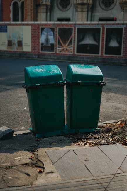 Two green wheelie bins with closed lids positioned side by side on a concrete sidewalk, with some dirt and debris around their base. The bins are placed near a curb, with a partially paved section beneath them. Behind the bins, a building with brick and stone facade features five boarded-up windows covered with painted wooden panels, and a circular decorative element is visible above. The lighting suggests an outdoor setting during daylight, with shadows cast on the ground. As part of professional cleaning and waste management practices, Cleaner Kingston upon Thames advises adherence to local council rules for waste disposal visible in the background, emphasizing proper sanitation and housekeeping in residential and commercial areas.