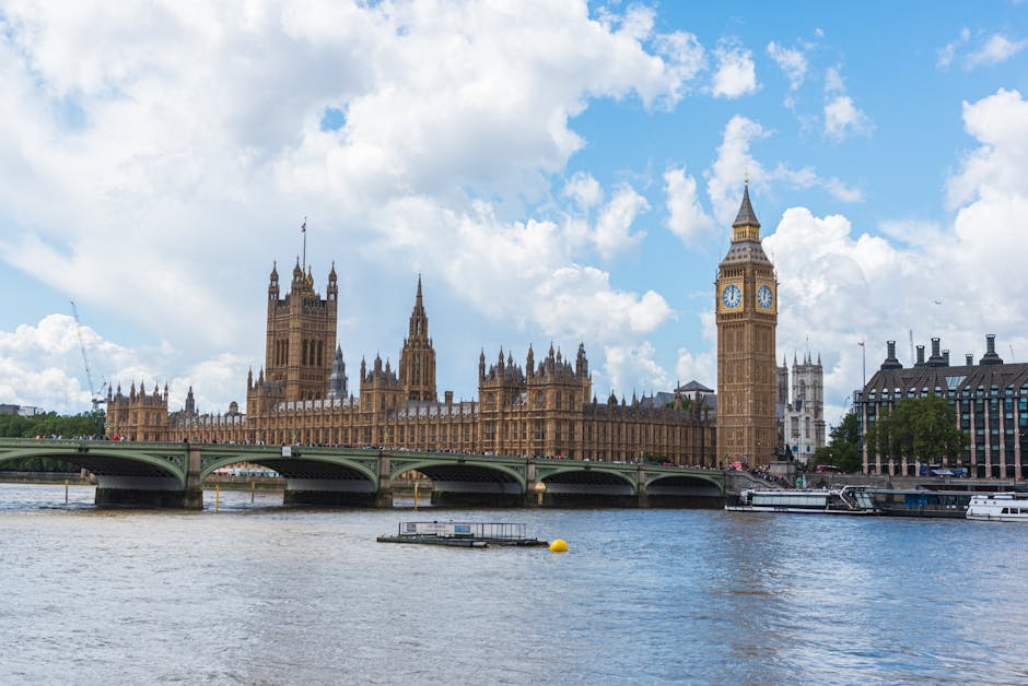 A panoramic view of the Palace of Westminster in London, featuring the iconic Big Ben clock tower with its stone facade, surrounded by a stone bridge spanning the River Thames. The river surface shows gentle ripples, with a few small boats moored along the sides. The sky is partly cloudy with bright, fluffy clouds illuminating the historic Gothic-style architecture, which appears well-maintained and clean. In the foreground, the water reflects the structures and sky, highlighting the clarity and calmness of the river. The overall scene emphasizes the urban landscape of central London, showcasing its architectural grandeur and cleanliness, consistent with the professional cleaning and maintenance services offered by Cleaner Kingston upon Thames.