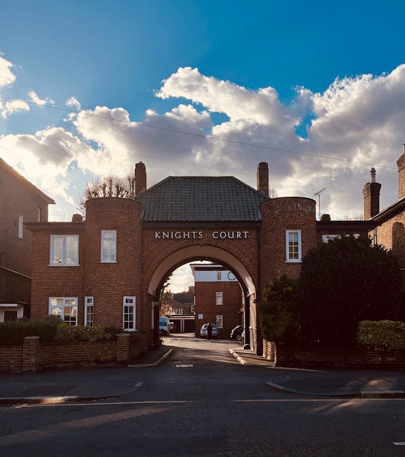 Photograph of the entrance to Knights Court, a brick residential building with a large central archway and the sign 'KNIGHTS COURT' above it, set against a partly cloudy sky with sunlight illuminating the scene. Surrounding the entrance are well-maintained gardens with neatly trimmed bushes, and a small road leading into the courtyard area. The building features white-framed windows and multiple chimneys on the roof, creating a classic London apartment appearance. This exterior view highlights the clean and tidy appearance of the property, consistent with the expert domestic cleaning, surface cleaning, and sanitisation services offered by Cleaner Kingston upon Thames, ensuring properties are maintained to high hygiene standards.